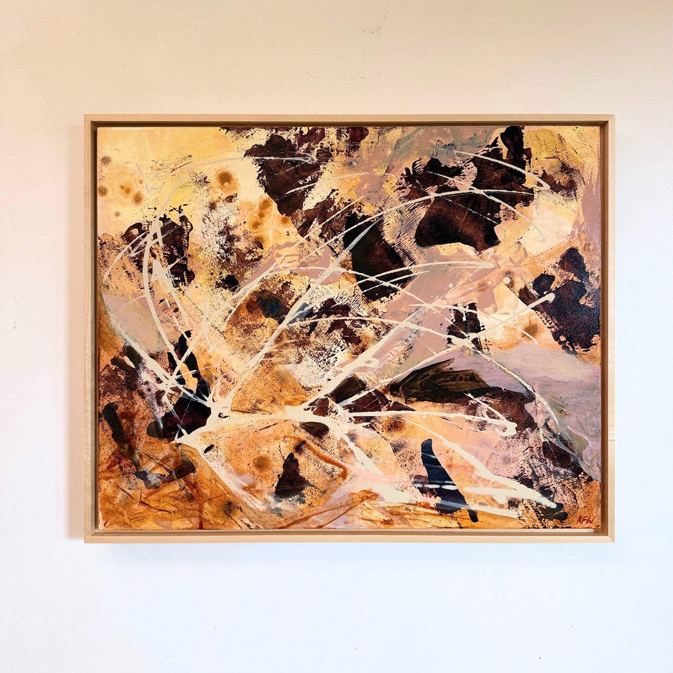 An abstract expressionist painting with splatters and brushstrokes in black, gold, beige, and white, housed in a light wood frame and mounted on a white wall.