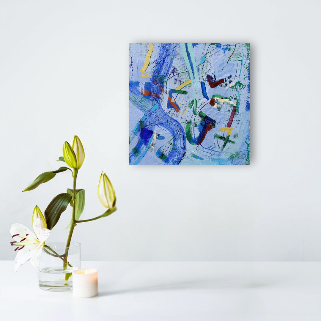 An abstract canvas painting with blue, green, yellow, and red brushstrokes hanging on a white wall above a white table. A clear glass vase with a white lily and a small white candle sit on the table.