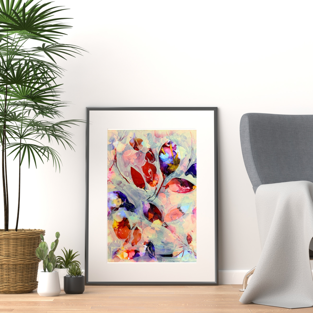 A framed abstract art print with colorful floral or leaf-like brushstrokes in red, orange, pink, purple, and blue tones, leaning against a white wall in a styled room with a potted plant, small cacti, and a gray chair.