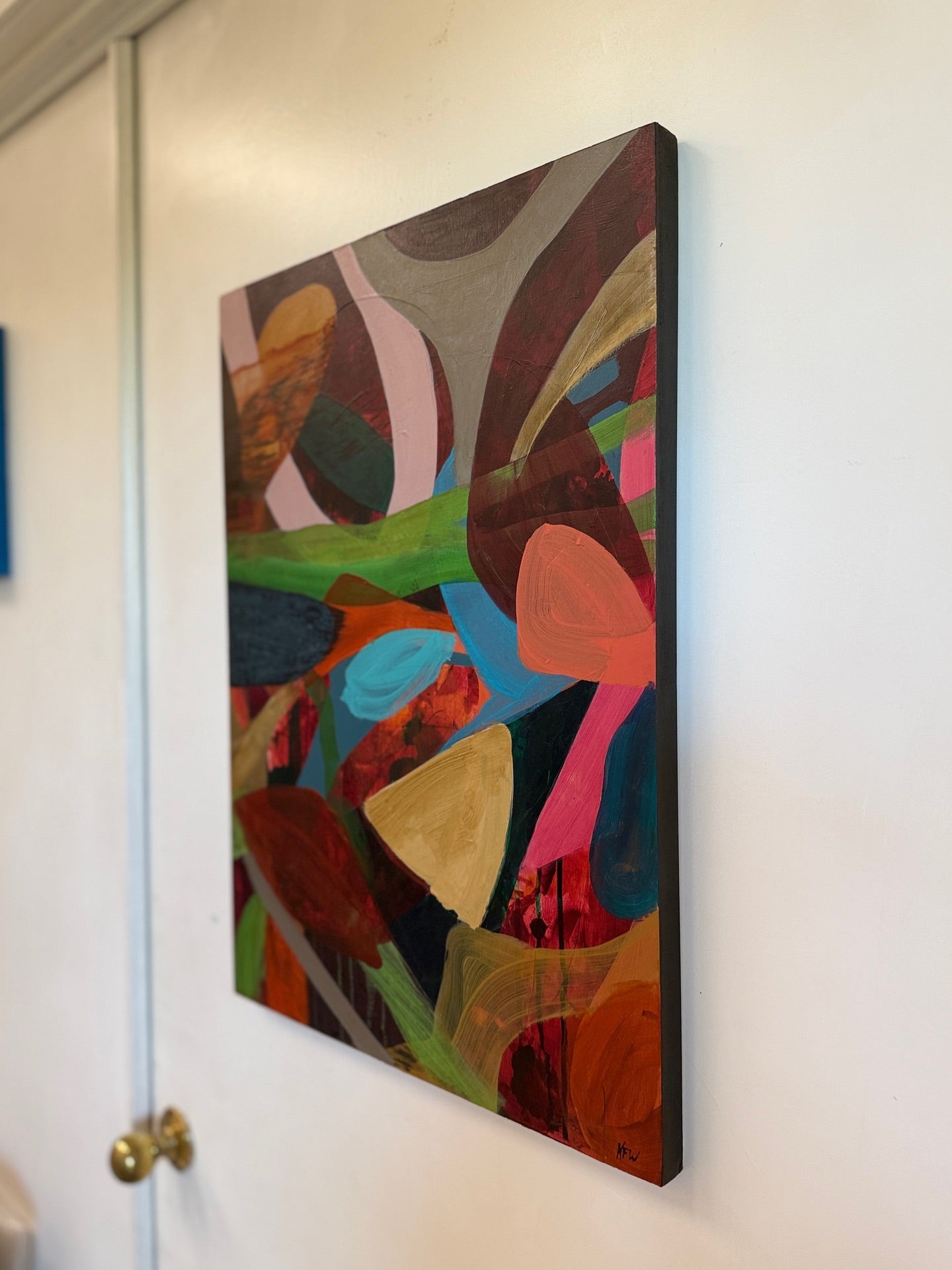 A side view of an abstract canvas painting with a colorful geometric pattern, hanging on a white wall next to a white door with a brass doorknob.