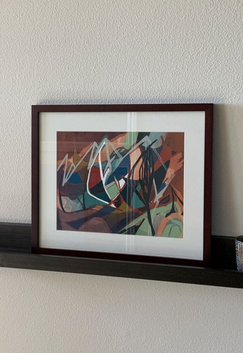 A framed abstract art print with a dark brown wood frame and white mat, hanging on a textured off-white wall above a dark shelf.