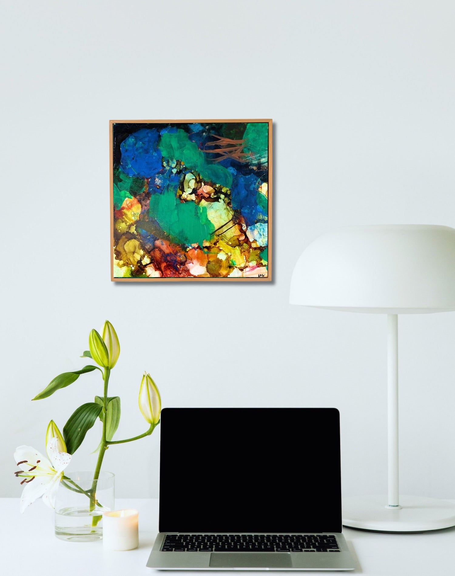Square abstract painting of blue, green and yellow in a white room above a laptop and flower stem.